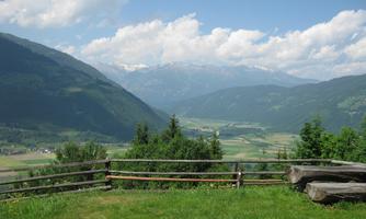 An idyllic mountain landscape with green meadows and majestic mountains in the background. The sky is blue and slightly cloudy.