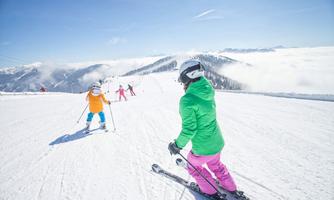 Skiers on a sunny slope with snow-covered mountains in the background. Children in colorful ski outfits enjoy the day in the snow.