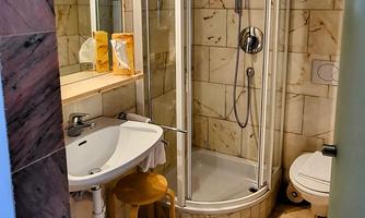 A bathroom with a shower, a sink, and a toilet. The walls are adorned with beige marble, and there is a small wooden bench.