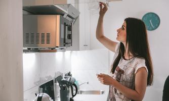 A woman is standing in a modern kitchen and hanging something on the wall. In the background, kitchen appliances and a clock are visible.