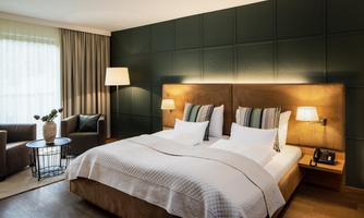 A modern hotel room with a large double bed and stylish furniture. The walls are dark green, and the room has plenty of natural light.