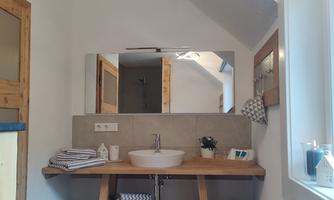 A modern bathroom with a wooden shelf and a large mirror. The decor is bright and friendly, adorned with towels and a small plant.