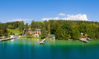 A tranquil landscape with a clear blue lake and lush green trees. On the opposite side, several houses and boats are visible.