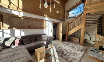 A cozy living room with wooden decor and large windows. The room contains a sofa, a table, and a staircase.