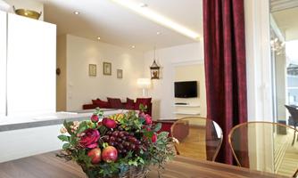 A beautiful table with a fruit basket stands in the foreground. In the background, you can see a cozy living room with red sofas and a TV setup.