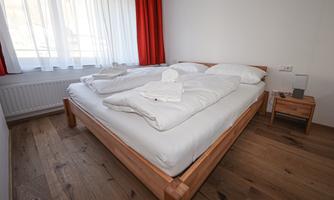 A bright bedroom with a double bed and fresh bedding. Large windows with curtains and a wooden floor create a cozy atmosphere.