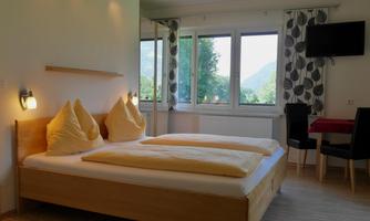 A bright room with a double bed, two pillows, and a large window overlooking the mountains. There is also a table and chairs as well as a television on the wall.