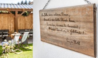 A rustic wooden sign with a handwritten message hangs on a wall. In the background, you can see a cozy garden area with seating.
