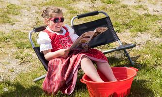 A girl in traditional dress sits relaxed in a chair and reads a magazine. Her feet are in a red bucket with water.