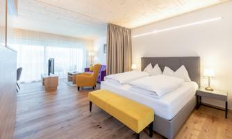 A modern hotel room with a large bed and comfortable seating areas. The room has a bright, inviting style and plenty of natural light.