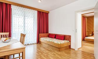 A bright living room with a cozy sofa and a dining area. Red curtains and wooden flooring create an inviting atmosphere.