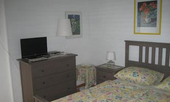 A simple bedroom with a bed, a dresser, and a television. The walls are light, and there are some decorative pictures.