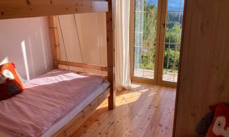 A cozy children's room with a bunk bed and a single bed. The windows offer a view of nature and plenty of daylight.
