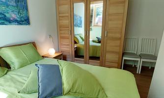 A cozy bedroom with a large bed and colorful pillows. In the background, there is a wooden wardrobe and two chairs.