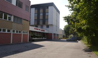 A modern building with several floors and a parking lot. In the background, trees can be seen and there is a sunny atmosphere.