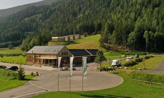 A modern building nestled in a picturesque landscape with green meadows and surrounding mountains. Several residential houses are visible in the background.