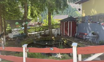 A cozy garden with a small pond and a red fence. In the background, trees and a few seating areas can be seen.