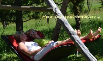 Two women are relaxing in a hammock under trees. The surroundings are green and sunny, perfect for enjoying.