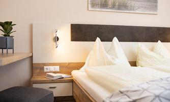 A cozy bedroom with a double bed and white pillows. On the table lies a book and in the background, there is a beautiful picture.