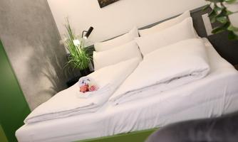 A cozy bedroom with a large bed and fresh white bed linens. Next to the bed is a small table with a plant and decorative flowers.