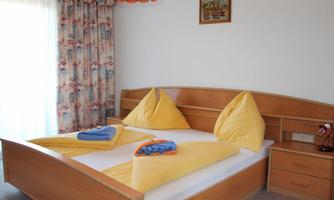 A bright bedroom with a large double bed and colorful pillows. The room has windows with curtains and a nightstand.