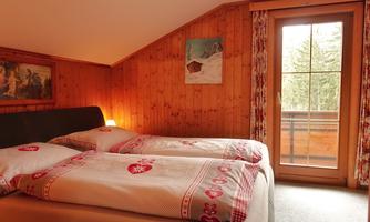 A cozy bedroom with wooden paneling and two single beds. Large windows offer a view of the balcony and the surrounding nature.