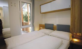 A modern bedroom with a double bed and a large window. There are wooden shelves and a mirror on the wall.