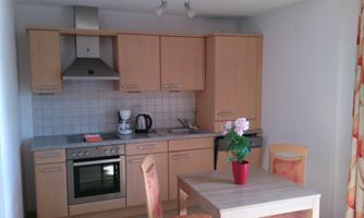 A modern kitchen with light wooden cabinets and a small table. On the table, there is a pot with a plant and some kitchen appliances are visible.