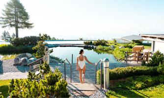 A beautiful outdoor area with a pond and lots of greenery. A woman in a swimsuit is walking towards a wooden terrace.