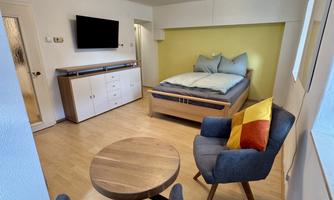 A cozy room with a double bed, a table, and two chairs. The walls are light, and there are large windows that provide daylight.