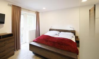 A modern bedroom with a double bed and a red bedspread. There is a television on the wall and large windows with curtains.