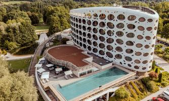 A modern hotel with a striking facade and round windows. The outdoor area features a pool and loungers in a beautiful green setting.