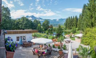 An inviting terrace with comfortable seating and sun umbrellas. In the background, the green forests and majestic mountains stretch under a blue sky.