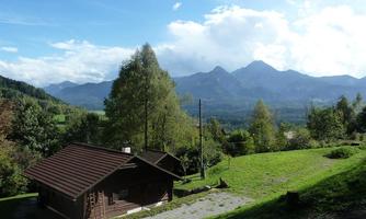 A cozy cabin surrounded by green meadows and trees. Majestic mountains rise in the background under a clear sky.