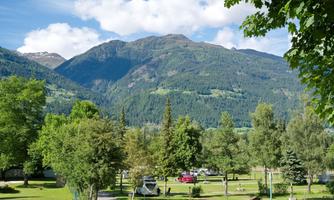 A green landscape with many trees and a view of big mountains. The sky is blue with some clouds.