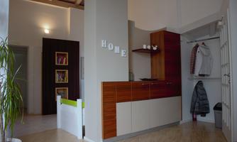 A modern entrance area with wooden cabinets and a decorative "HOME" sign. Jackets hang on the wall, and there are plants and bookshelves nearby.