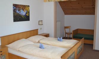 A cozy bedroom with two single beds and a wooden wardrobe. In the background, there is a small seating area with a chair and a bench.