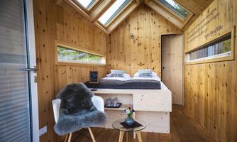 A cozy, modern wooden house with large windows and plenty of natural light. The interior is designed in a minimalist style with a bed, a table, and a chair.