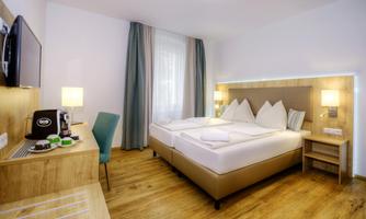 A modern hotel room with two beds and wooden flooring. The walls are decorated in light colors, and there are windows with curtains as well as a television.