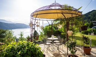 A cozy pavilion with a table and chairs is set in a blooming garden. In the background, there is a beautiful view of a lake and the surrounding mountains.