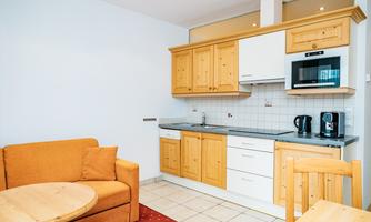 A modern kitchen with wooden cabinets and gray countertops. In the foreground are a round table and an orange sofa.