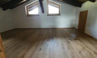 An empty room with wooden floors and sloping ceiling beams. The windows let in a lot of daylight.