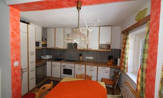 A modern kitchen with an orange accent wall and a bright decor. The table is set with an orange tablecloth and there are colorful curtains.