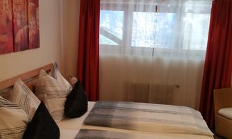 A cozy bedroom with two beds and elegant pillows. Large windows let in plenty of light, and the curtains give the room a warm atmosphere.