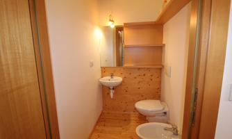 A small bathroom with wooden shelves and sanitary facilities. The walls are bright and the floor is made of wood.