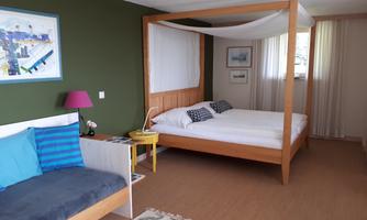 A modern room with a four-poster bed and stylish furniture. The walls are painted in a soft green shade, and plenty of daylight pours through the curtains.