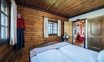 A cozy bedroom with wood paneling and a double bed. Next to the window hangs a traditional garment, and in the background, another room is visible.