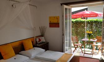 A bright room with two beds and yellow cushions. Through the open door, a small table is visible outside with chairs and an umbrella.