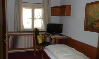 A simple room with a single bed, a desk, and a television. The windows let in plenty of light and offer a view of the nature outside.