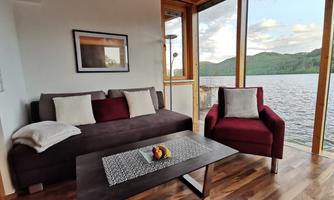 A cozy living room with a sofa and an armchair, overlooking a lake and the surrounding mountains. Large windows let in plenty of daylight.
